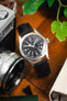 Close-up of a Hamilton field watch with a black dial and white Arabic numerals on a black leather strap, placed on a wooden surface beside a vintage camera lens, notebook, and green foliage.