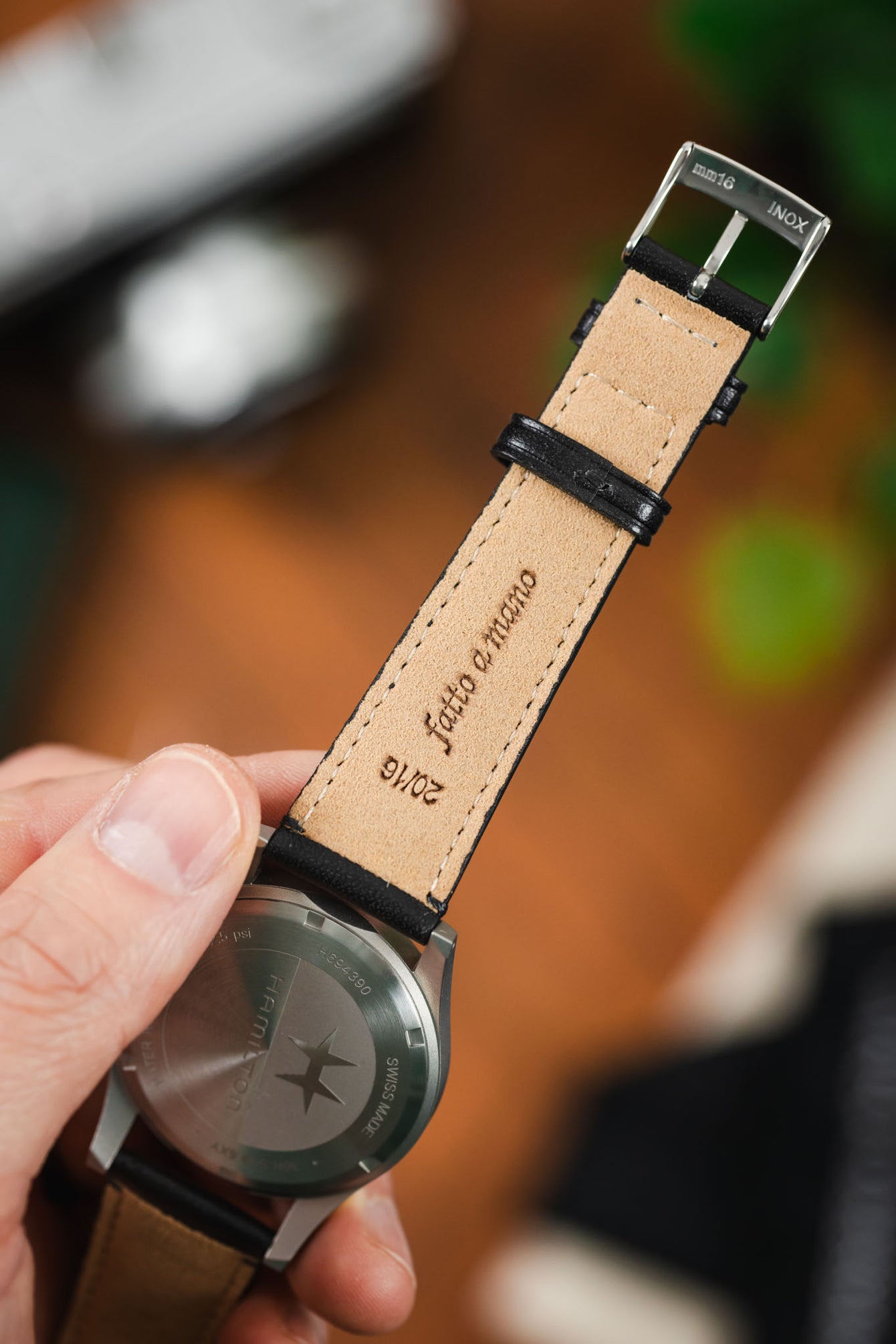 Close-up of the underside of a black leather watch strap with beige lining, showing “Fatto a Mano” and “20/16” markings, held beside a brushed steel watch caseback.