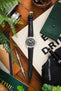 Flat-lay of a Hamilton field watch with a black dial and black leather strap, arranged on a wooden surface with a camera, screwdriver, notebook, greenery, and a drink, creating a lifestyle workspace scene.