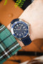 Close-up of an Omega Seamaster diver’s watch with a blue dial and bezel, worn on a wrist with a navy leather strap and a green plaid sleeve visible.