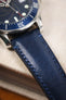 Macro close-up of a navy leather watch strap with tonal stitching attached to a brushed steel diver’s watch case, highlighting the smooth grain, stitching detail, and bezel edge against a textured fabric surface.