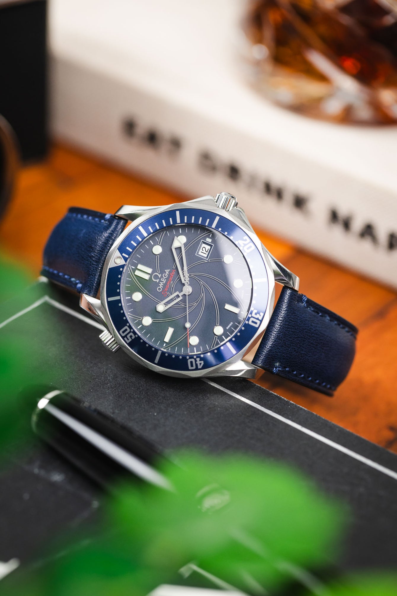Close-up of an Omega Seamaster diver’s watch with a blue dial and bezel on a navy leather strap, resting on a dark surface with blurred greenery and a whisky glass in the background.