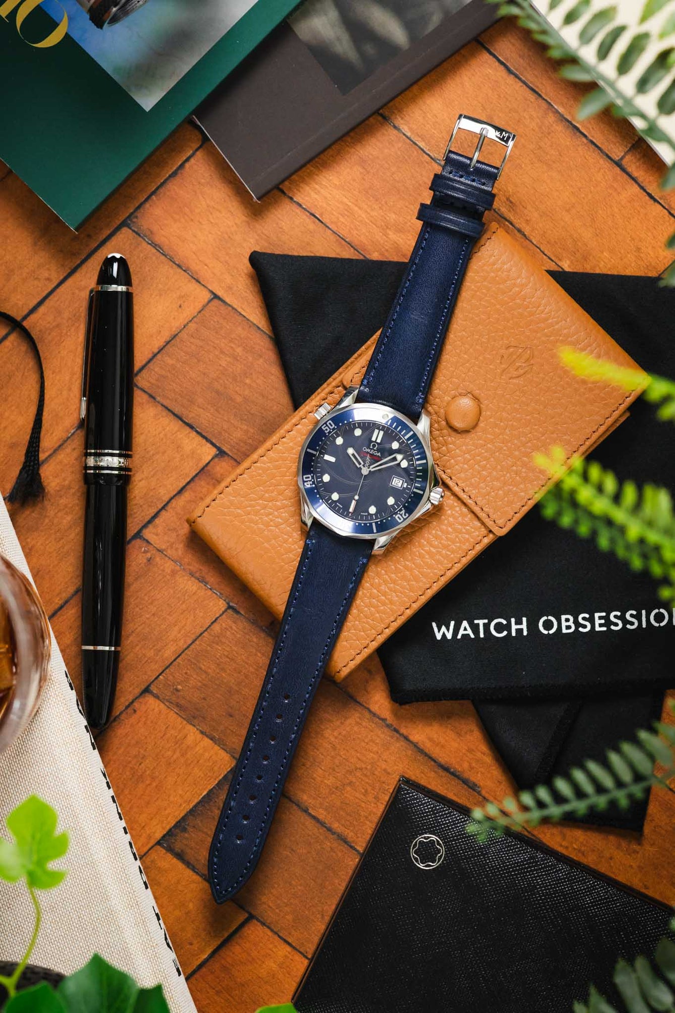 Flat-lay of an Omega diver’s watch with a blue dial on a navy leather strap, placed on a tan leather case and surrounded by a pen, pouch, greenery, and desk accessories on a wooden surface.