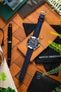 Flat-lay of an Omega diver’s watch with a blue dial on a navy leather strap, placed on a tan leather case and surrounded by a pen, pouch, greenery, and desk accessories on a wooden surface.