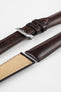 Brown leather watch strap with contrast stitching, shown in angled close-up with a silver buckle, adjustment holes, and beige suede underside.