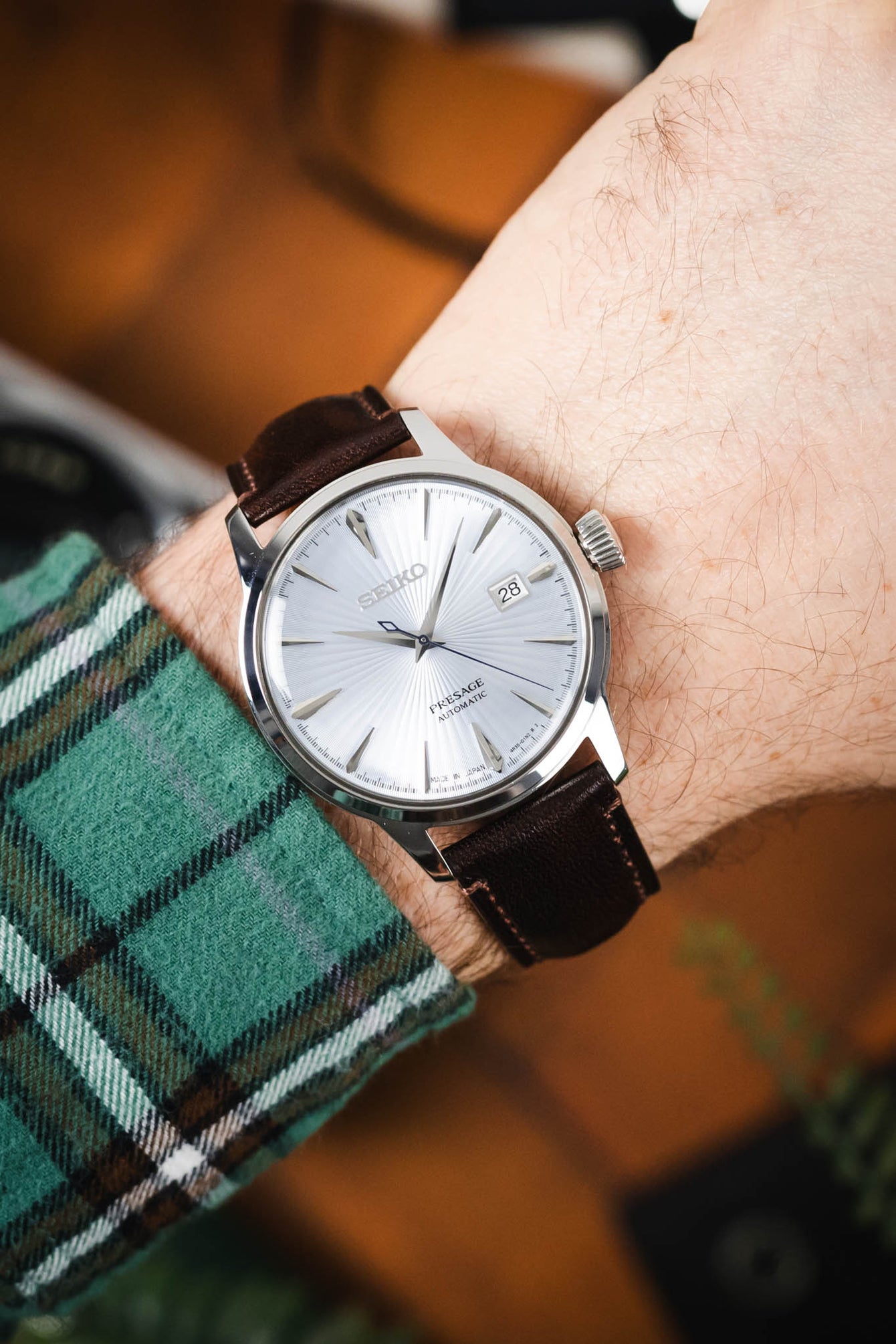 Close-up of a Seiko Presage watch with a textured white dial and date window, worn on a brown leather strap with contrast stitching, paired with a green plaid sleeve.