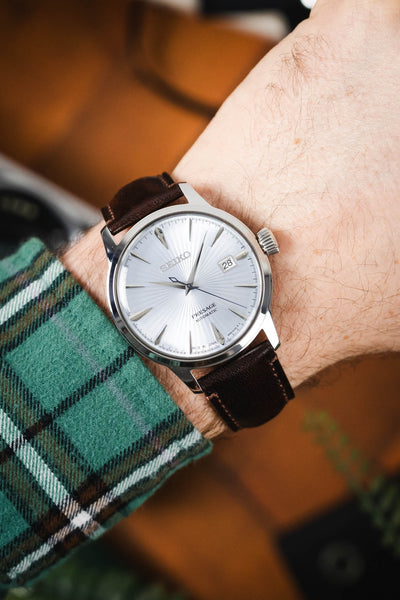 Close-up of a Seiko Presage watch with a textured white dial and date window, worn on a brown leather strap with contrast stitching, paired with a green plaid sleeve.