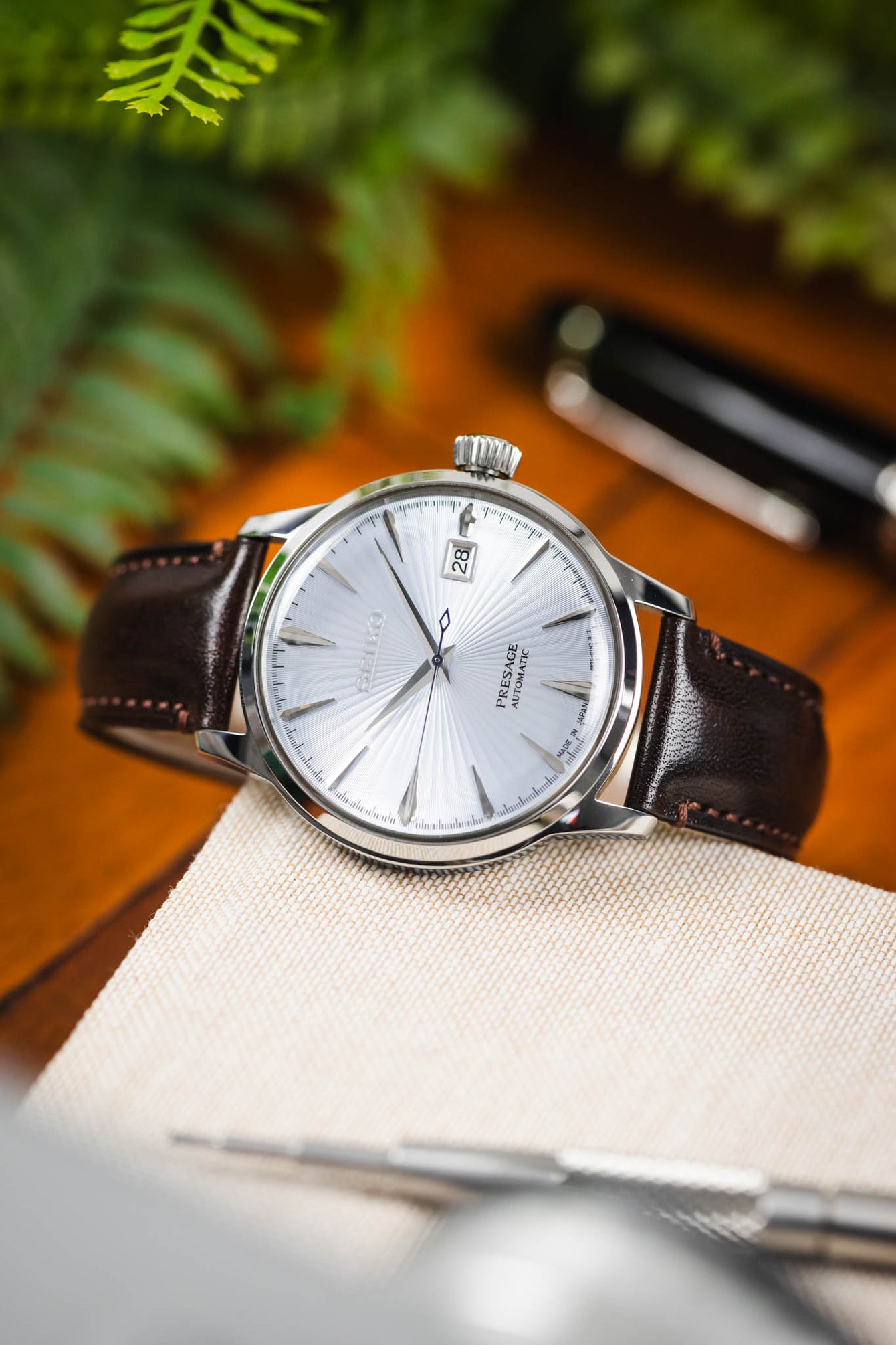 Close-up of a Seiko Presage watch with a textured white dial and date display, paired with a glossy brown leather strap. The watch rests on a light fabric surface, with greenery and a pen softly blurred in the background.