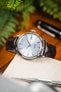 Close-up of a Seiko Presage watch with a textured white dial and date display, paired with a glossy brown leather strap. The watch rests on a light fabric surface, with greenery and a pen softly blurred in the background.