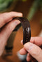 Macro close-up of a brown leather watch strap with a textured finish and contrast stitching, held between fingers, highlighting adjustment holes and the glossy surface against a softly blurred background.