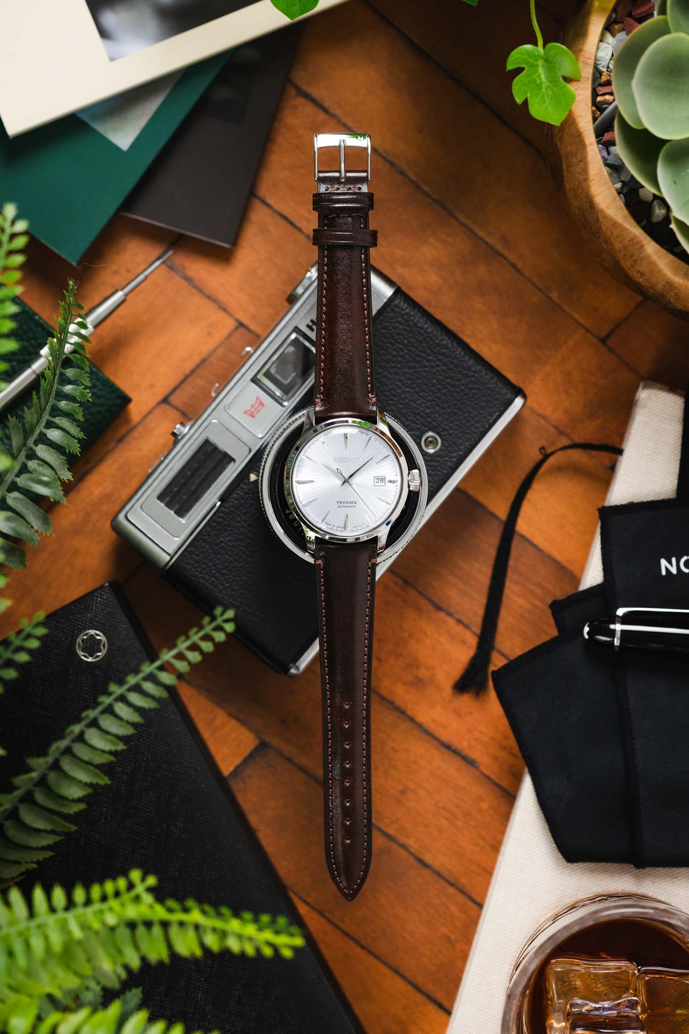 Flat-lay of a Seiko Presage watch with a white textured dial on a brown leather strap, placed over a vintage camera and surrounded by greenery, stationery, and accessories on a wooden surface.