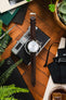 Flat-lay of a Seiko Presage watch with a white textured dial on a brown leather strap, placed over a vintage camera and surrounded by greenery, stationery, and accessories on a wooden surface.