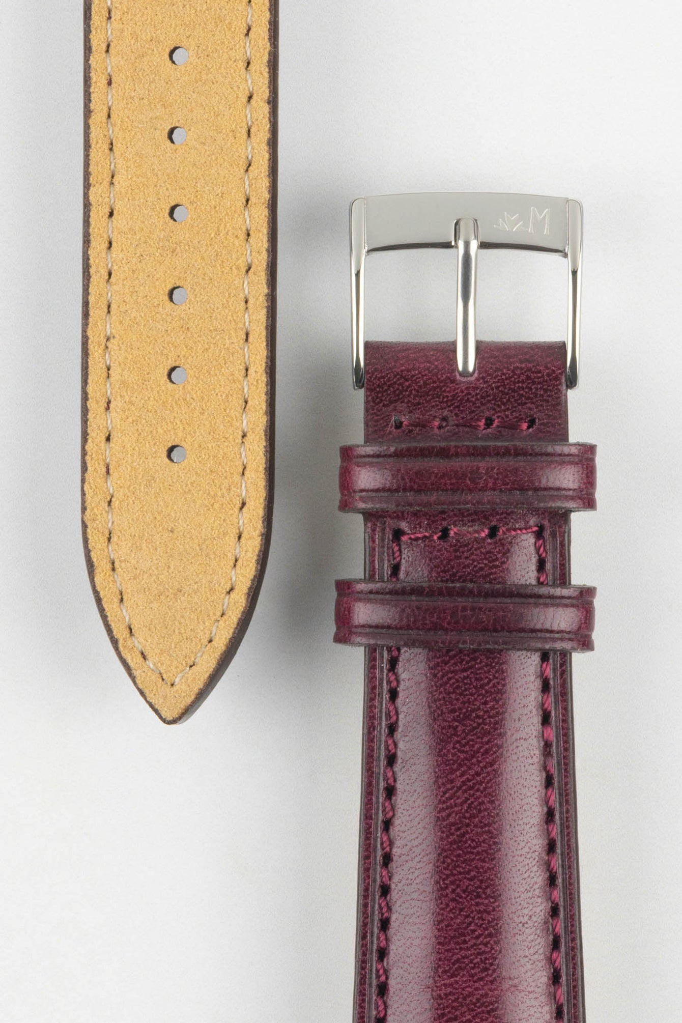 Close-up product shot of a burgundy leather watch strap and a separate strap piece displayed side by side on a plain light background. The strap features a smooth, slightly textured finish with matching burgundy stitching and multiple adjustment holes. The main strap is fitted with a polished silver pin buckle engraved with a small logo, while the reverse side of the separate piece reveals a tan suede underside with light contrast stitching, highlighting the materials, craftsmanship, and hardware details.
