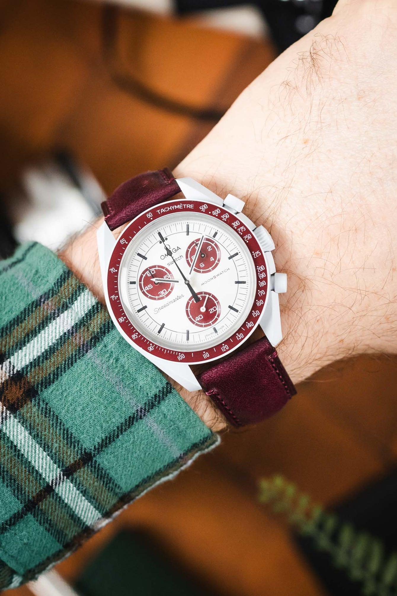 Close-up of an Omega x Swatch MoonSwatch with a white dial and burgundy bezel, worn on a wrist with a matching burgundy leather strap and a green plaid sleeve visible.
