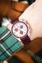 Close-up of an Omega x Swatch MoonSwatch with a white dial and burgundy bezel, worn on a wrist with a matching burgundy leather strap and a green plaid sleeve visible.