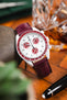 Close-up of an Omega x Swatch MoonSwatch with a white dial and burgundy bezel on a matching burgundy leather strap, styled beside a glass of whisky and a fountain pen on a wooden surface.