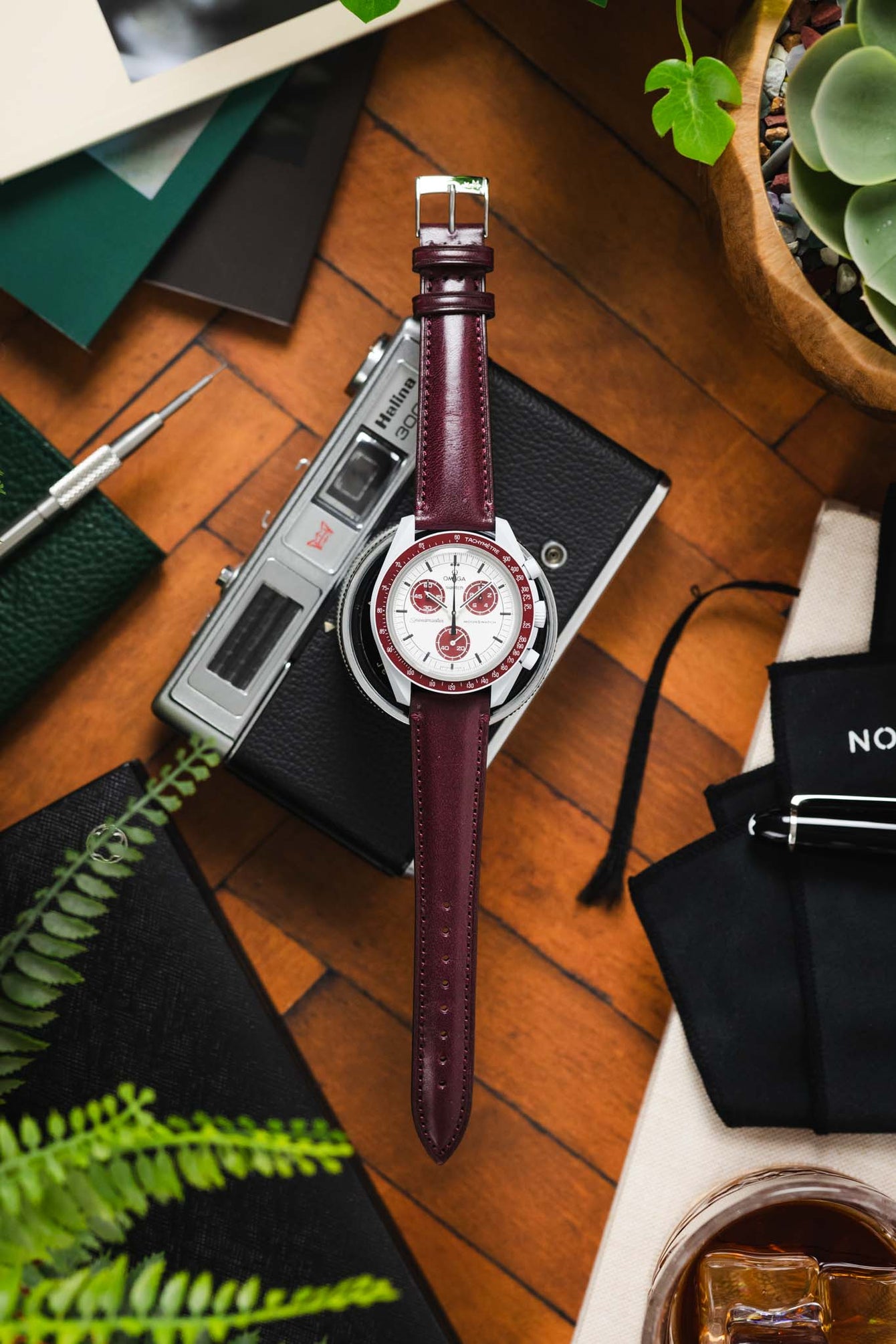 Flat-lay of an Omega x Swatch MoonSwatch with a white dial and burgundy bezel on a matching burgundy leather strap, arranged over a camera and surrounded by a screwdriver, pouch, greenery, and a glass of whisky on a wooden surface.