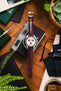 Flat-lay of an Omega x Swatch MoonSwatch with a white dial and burgundy bezel on a matching burgundy leather strap, arranged over a camera and surrounded by a screwdriver, pouch, greenery, and a glass of whisky on a wooden surface.