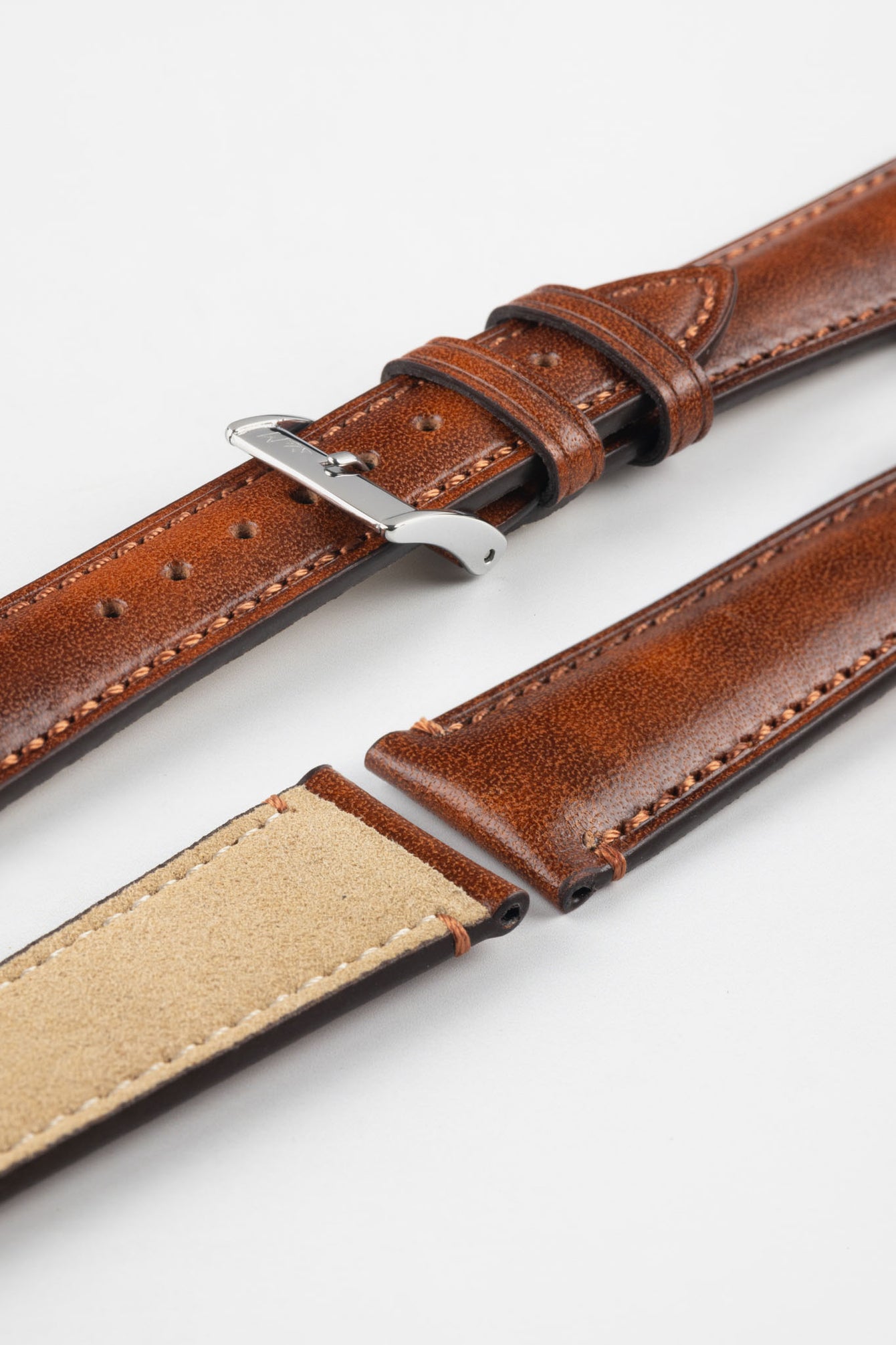 Tan-brown leather watch strap with matching stitching, shown in angled close-up with a silver buckle, adjustment holes, and beige suede underside.