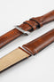 Tan-brown leather watch strap with matching stitching, shown in angled close-up with a silver buckle, adjustment holes, and beige suede underside.