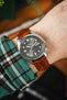 Close-up of a Seiko automatic watch with a grey dial and day-date display, worn on a wrist with a warm brown leather strap and green plaid sleeve visible.