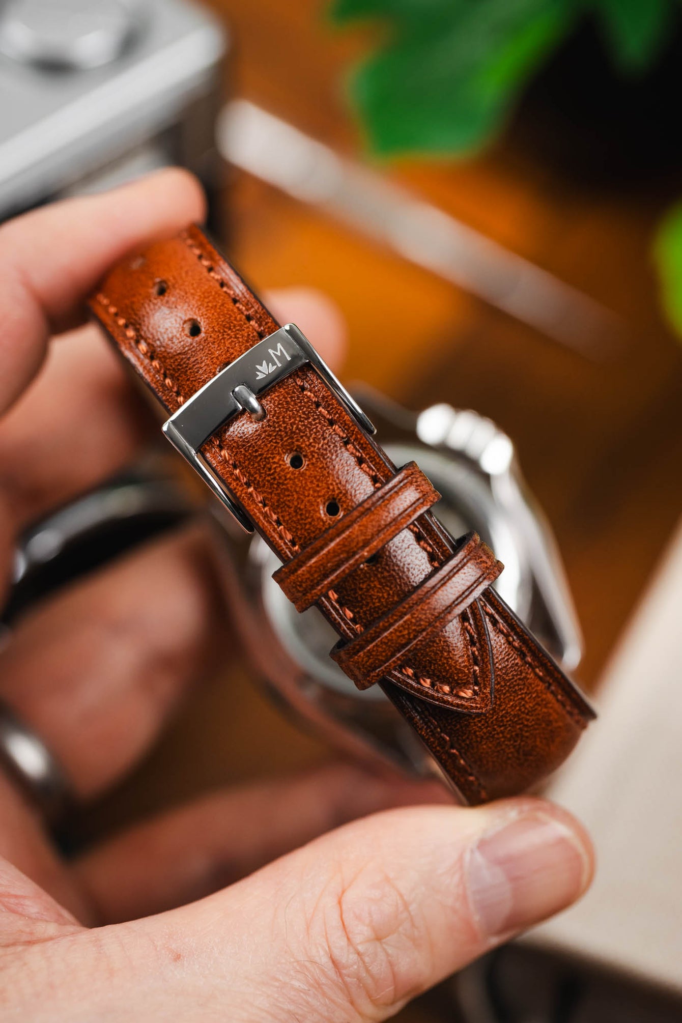 Macro close-up of a brown leather watch strap with a polished silver buckle engraved with an “M” logo. The strap features a smooth, slightly glossy finish, contrast stitching, multiple adjustment holes, and two keeper loops, held between fingers with a softly blurred background of a watch case and warm-toned setting.
