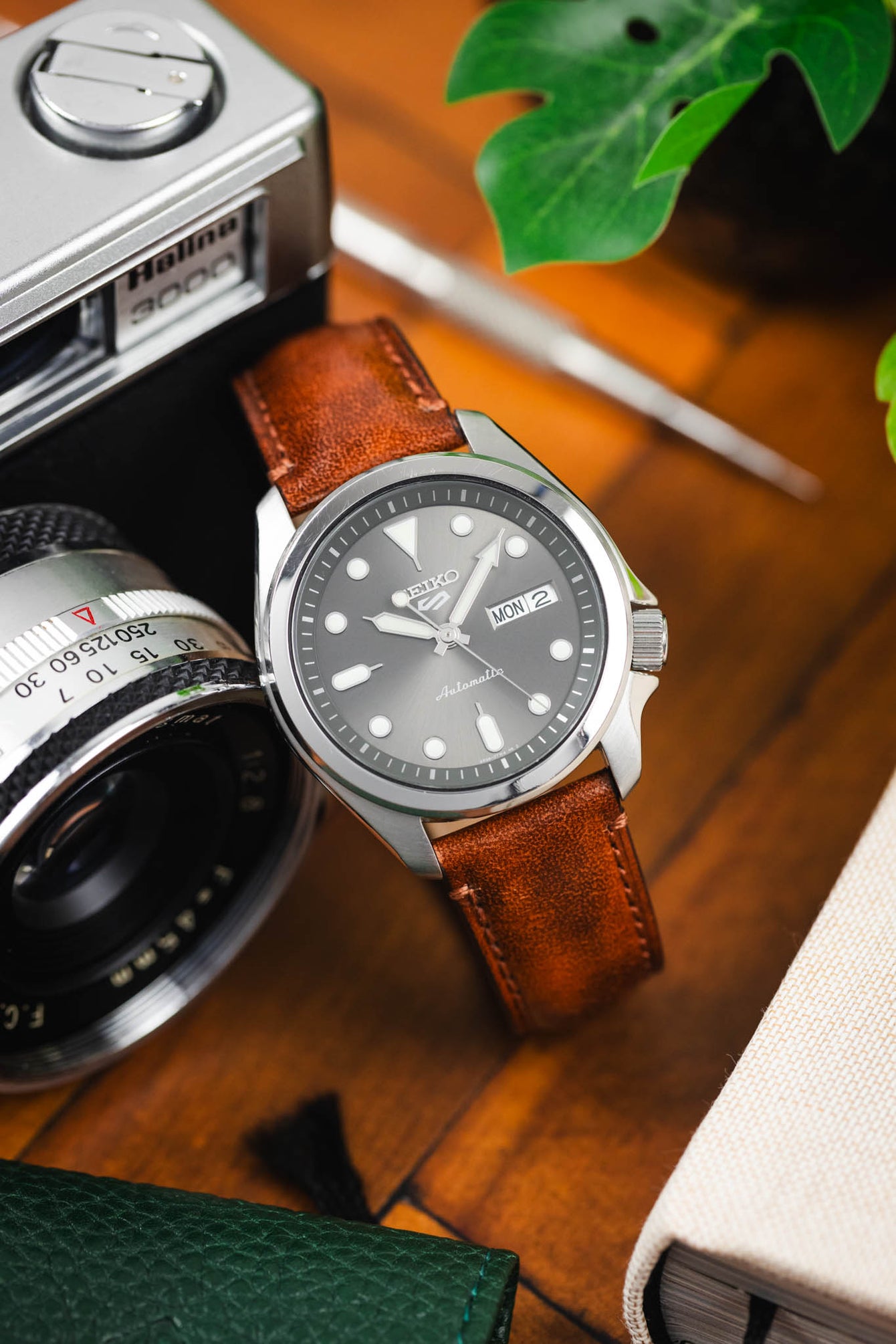 Close-up of a Seiko automatic watch with a grey dial and day-date window, paired with a brown leather strap, styled beside a vintage camera, notebook, and greenery on a wooden surface.