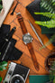 Flat-lay of a Seiko automatic watch on a warm brown leather strap, arranged on a wooden surface with a vintage camera, screwdriver, pen, cloth pouch, greenery, and a glass of whisky.