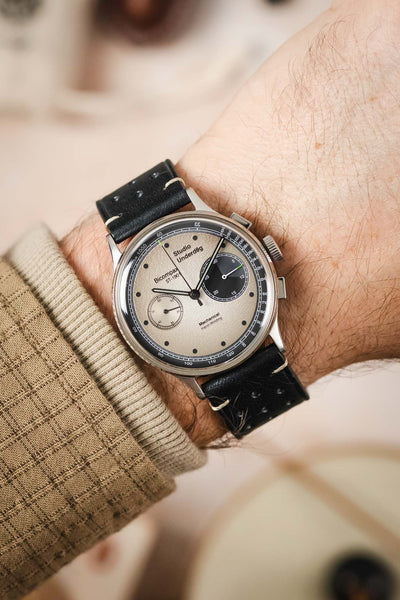 
A high-angle shot of a man's wrist, wearing a vintage-style chronograph watch with a light-coloured dial on a black Morellato Brahms perforated leather strap. The watch has two sub-dials and the man is wearing a tan sweater.