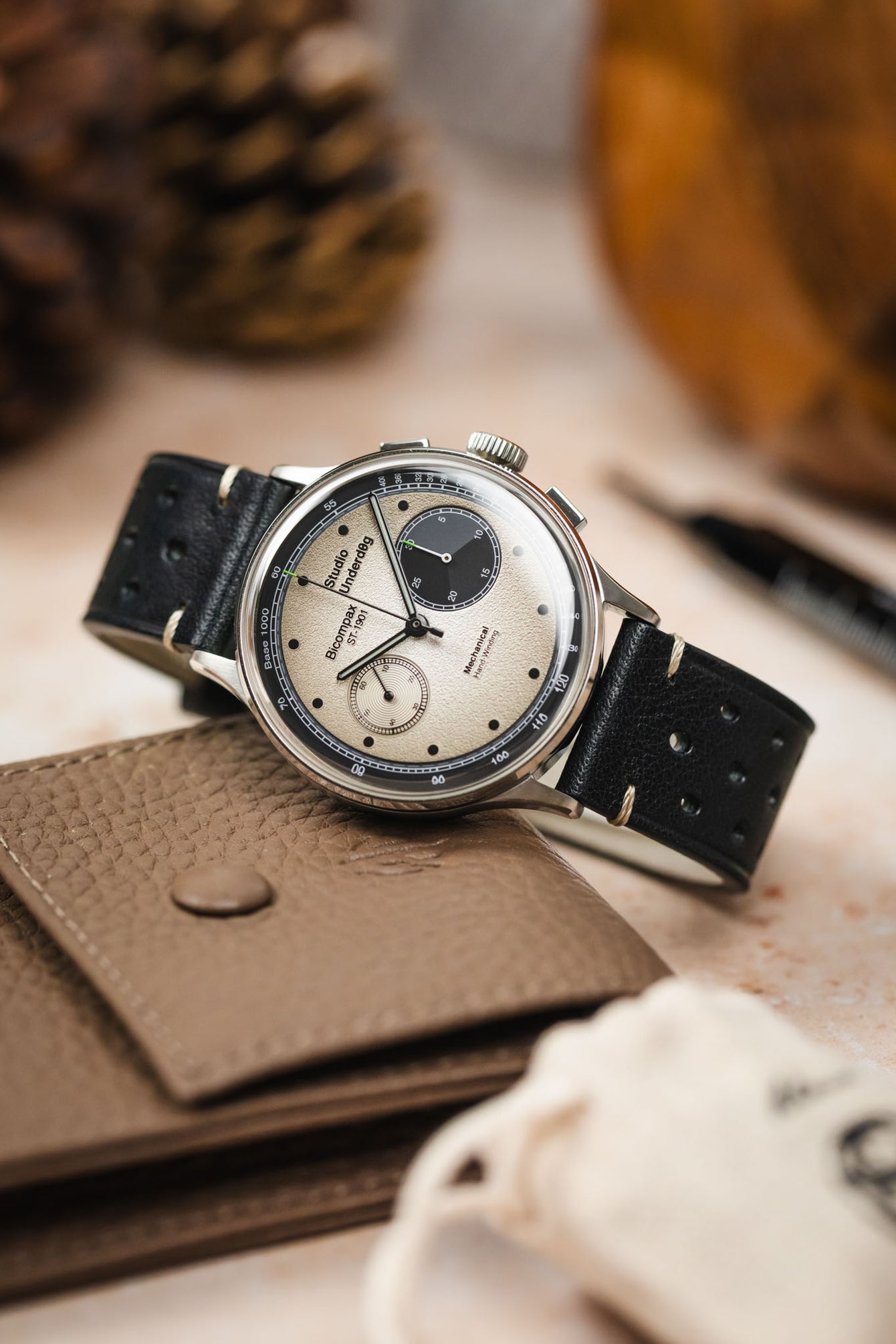 A vintage-style chronograph watch with a light-coloured dial and a black Morellato Brahms perforated leather strap, sitting on top of a tan leather wallet. In the soft-focus background are pine cones and a watch tool.
