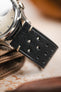 
A macro shot of a black Morellato Brahms watch strap, attached to a watch case, with a focus on the perforations and the white stitching at the lug ends. The watch is resting on a textured, light brown surface.