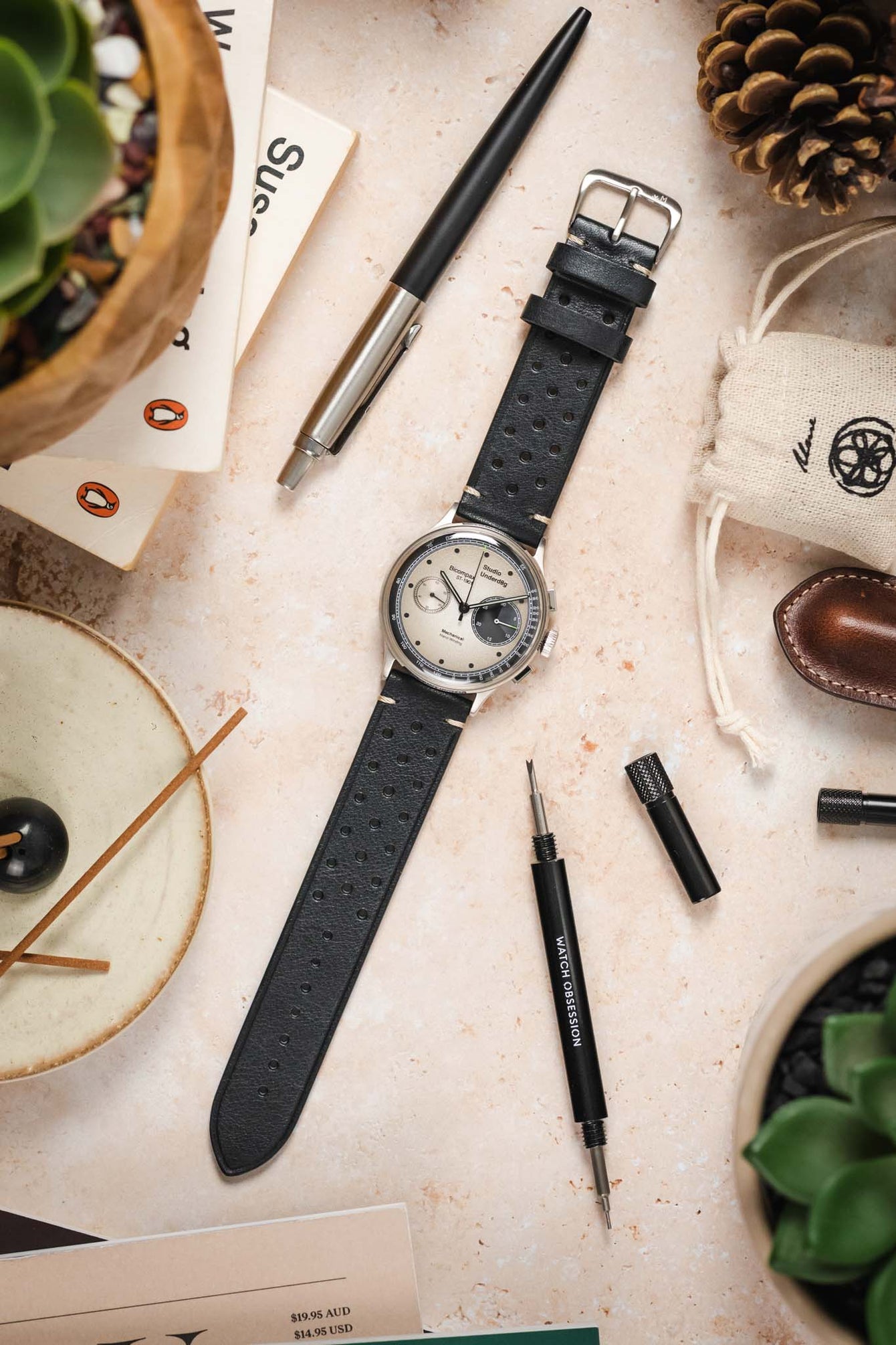 A flat-lay shot of a vintage-style watch with a light dial and a black Morellato Brahms perforated watch strap. The watch is surrounded by various objects, including a pen, small potted plants, a small linen bag, and watch tools, all arranged on a light-coloured, textured surface.