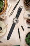A flat-lay shot of a vintage-style watch with a light dial and a black Morellato Brahms perforated watch strap. The watch is surrounded by various objects, including a pen, small potted plants, a small linen bag, and watch tools, all arranged on a light-coloured, textured surface.
