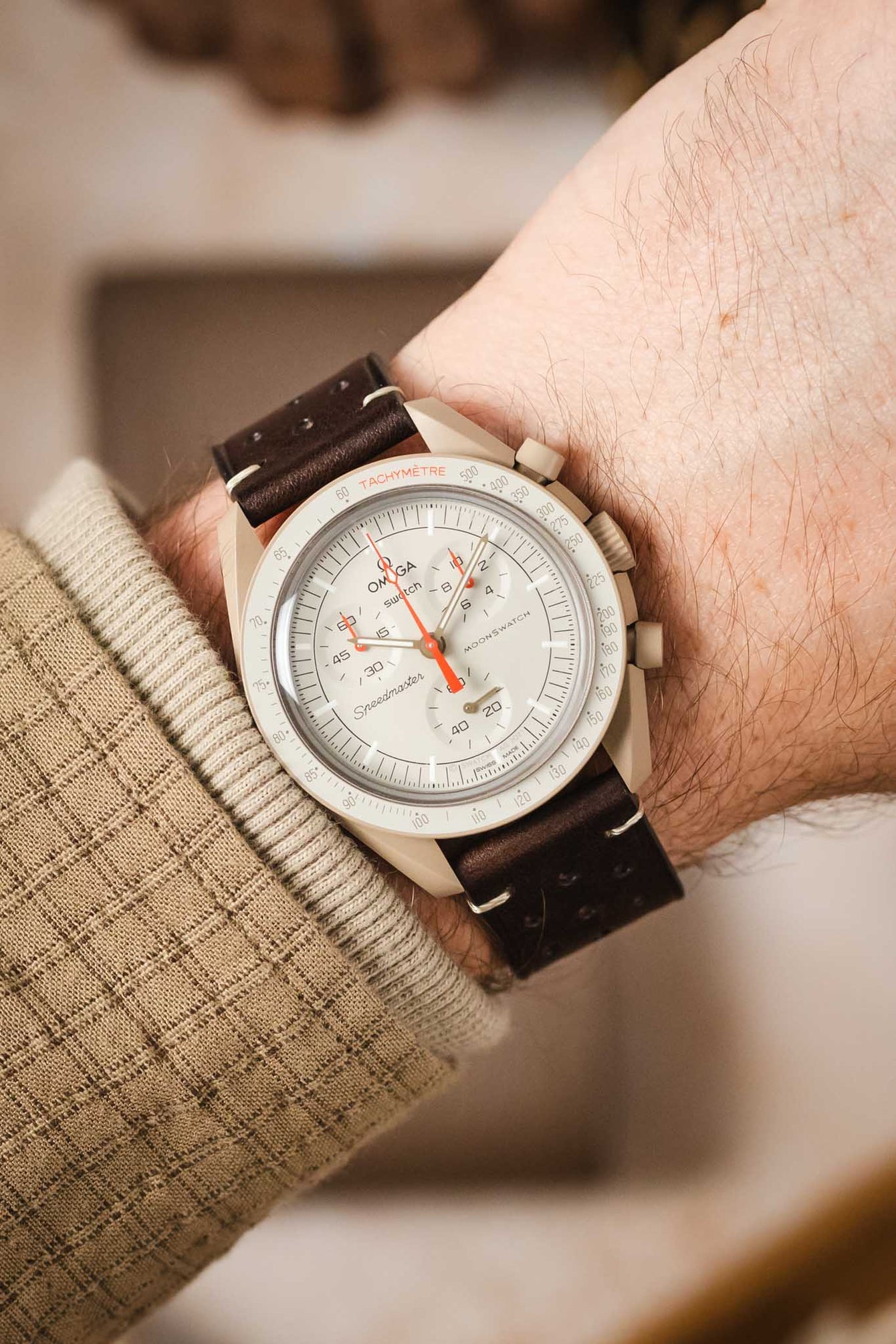 A high-angle shot of a man's wrist wearing an Omega x Swatch MoonSwatch 'Moon' watch on a dark brown Morellato Brahms leather strap. The watch has a white dial, white tachymeter bezel, and red hands. The man is wearing a tan sweater.