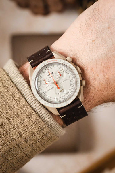 A high-angle shot of a man's wrist wearing an Omega x Swatch MoonSwatch 'Moon' watch on a dark brown Morellato Brahms leather strap. The watch has a white dial, white tachymeter bezel, and red hands. The man is wearing a tan sweater.