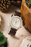
An Omega x Swatch MoonSwatch 'Moon' watch on a dark brown Morellato Brahms perforated leather strap, sitting next to a wooden bowl. In the soft-focus background are pine cones and a potted succulent.
