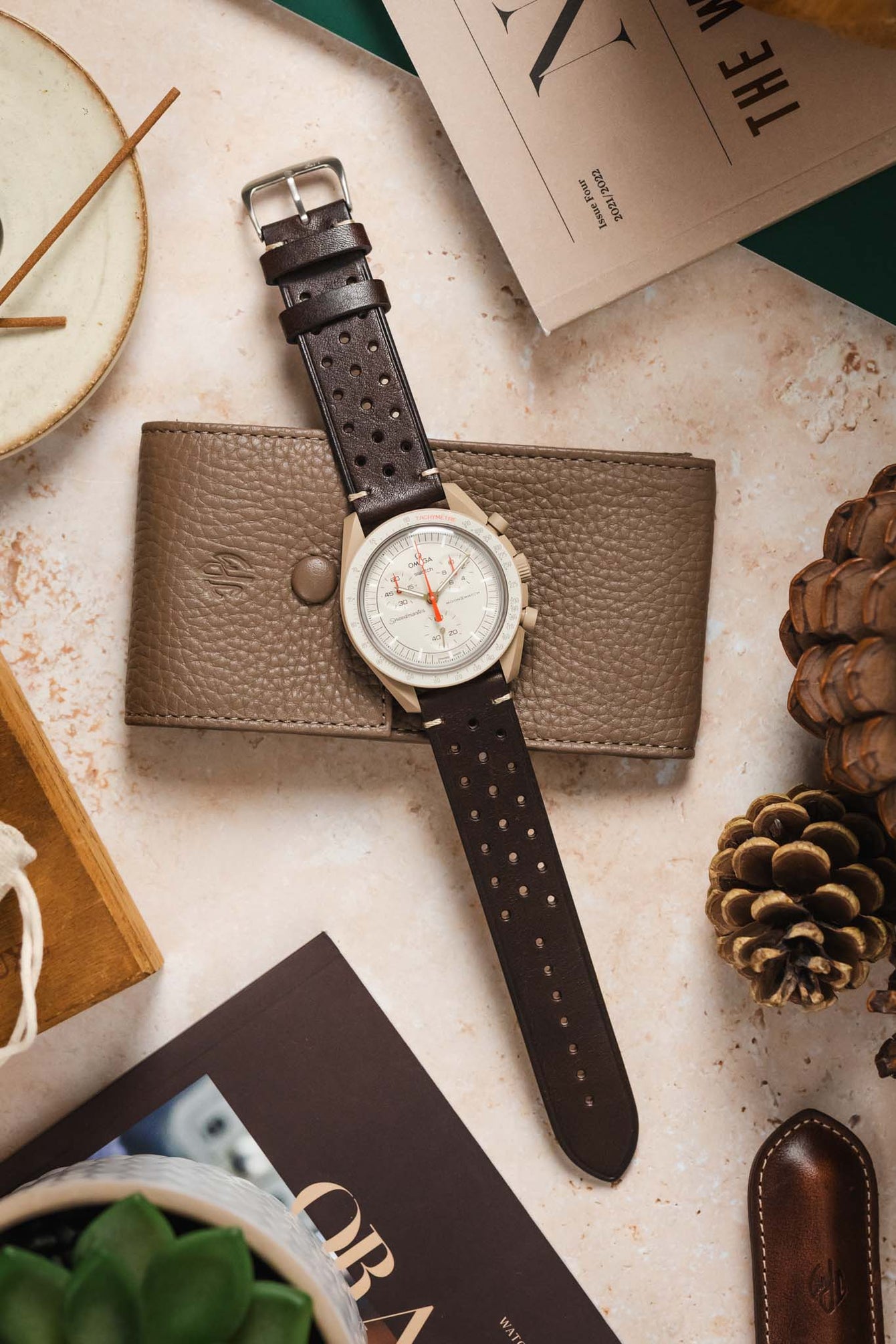 
A flat-lay shot of an Omega x Swatch MoonSwatch 'Jupiter' watch with a dark brown Morellato Brahms perforated watch strap. The watch is placed on a tan leather wallet and surrounded by a variety of items, including a magazine, pine cones, and a potted succulent.