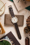 
A flat-lay shot of an Omega x Swatch MoonSwatch 'Jupiter' watch with a dark brown Morellato Brahms perforated watch strap. The watch is placed on a tan leather wallet and surrounded by a variety of items, including a magazine, pine cones, and a potted succulent.
