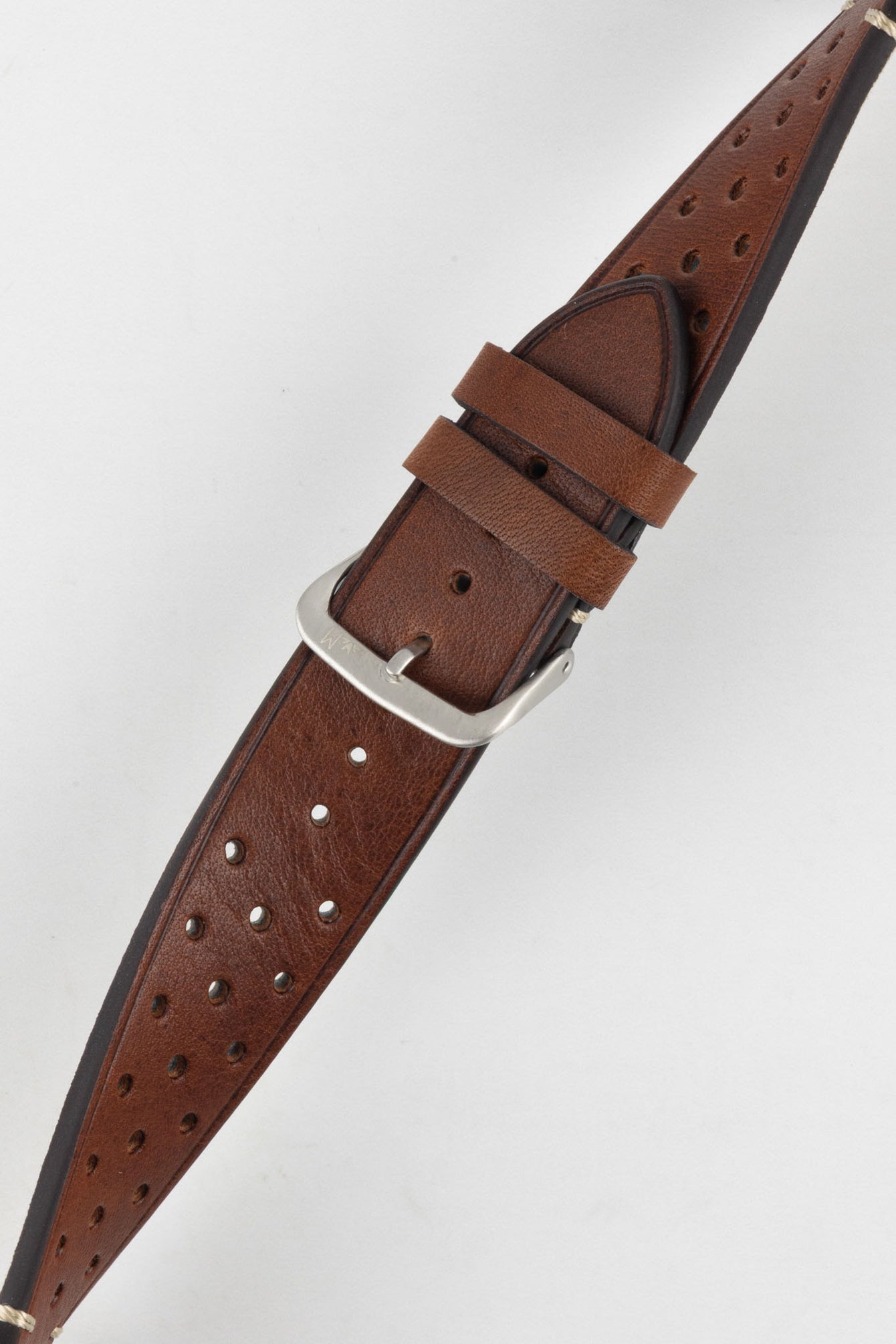 
A close-up, high-angle shot of a medium brown, 'gold' Morellato Brahms watch strap, buckled and resting diagonally against a white background. The image focuses on the rally-style perforations and the satin-finished steel buckle.