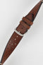 
A close-up, high-angle shot of a medium brown, 'gold' Morellato Brahms watch strap, buckled and resting diagonally against a white background. The image focuses on the rally-style perforations and the satin-finished steel buckle.