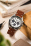 
A Seiko Speedtimer chronograph watch with a white dial and a medium brown Morellato Brahms perforated leather strap, sitting on a light-coloured wooden box. In the soft-focus background are books and a watch tool.