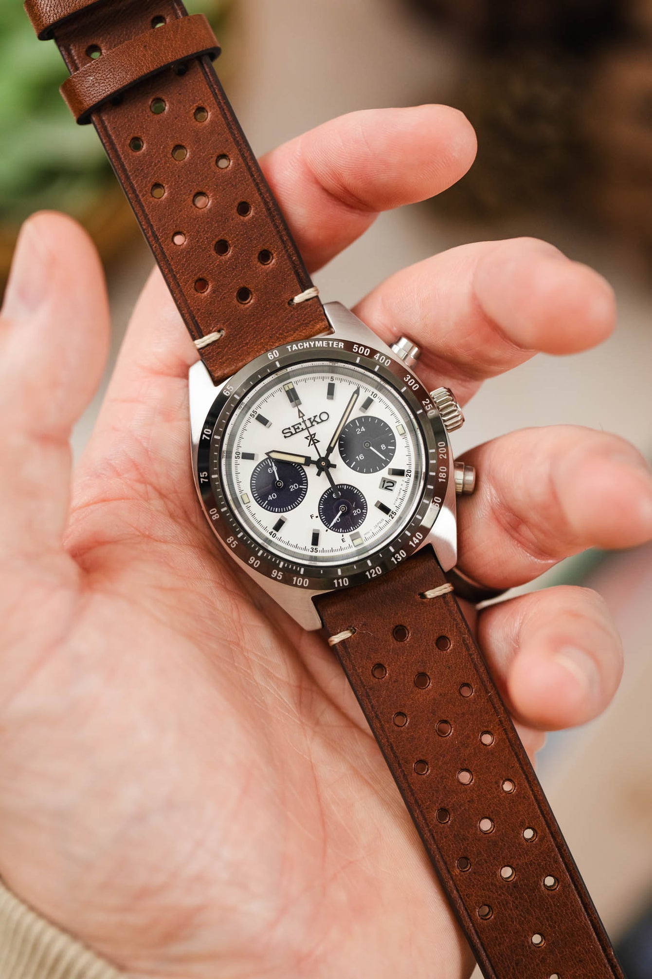 
A close-up shot of a hand holding a Seiko Speedtimer chronograph watch. The watch has a white dial with three black sub-dials and a black bezel, and it's on a medium brown Morellato Brahms perforated leather strap with beige stitching.