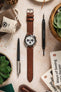 
A flat-lay shot of a Seiko Speedtimer chronograph watch with a medium brown Morellato Brahms perforated watch strap. The watch is surrounded by various objects, including a pen, books, a small linen bag, and watch tools, all arranged on a light-coloured, textured surface.