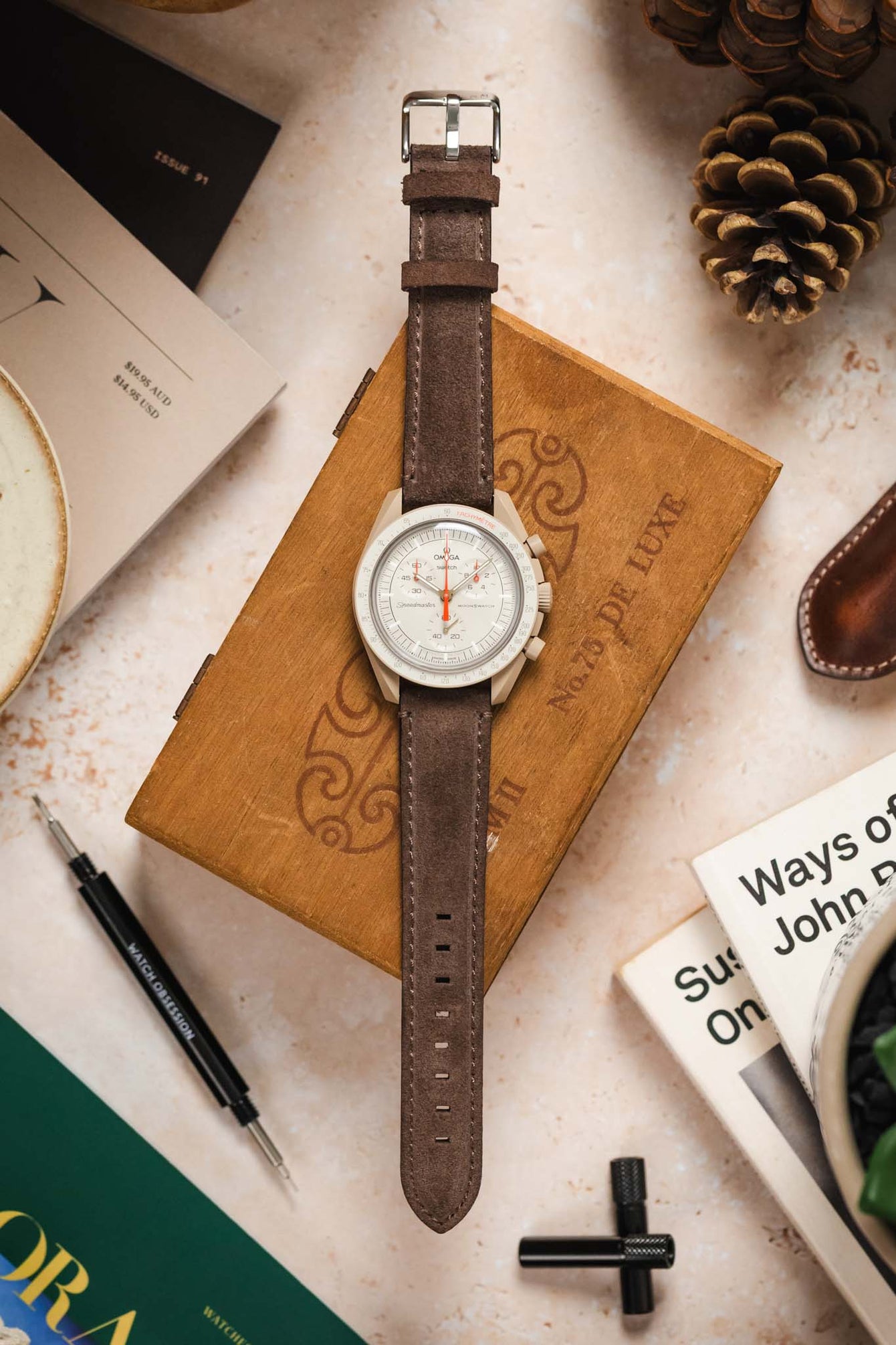 Overhead shot of a Morellato SCHUMANN brown suede watch strap with an Omega MoonSwatch and other accessories.