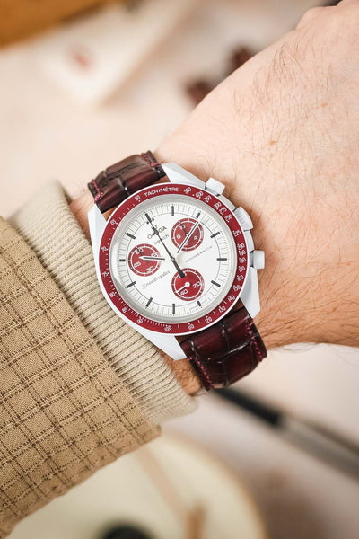A person's wrist wearing an Omega MoonSwatch with a stunning burgundy Morellato Tiepolo watch strap.