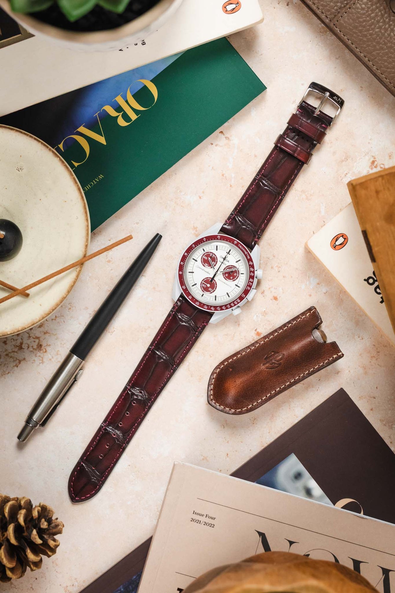 A burgundy Morellato Tiepolo watch strap attached to an Omega MoonSwatch, photographed with other watch accessories.
