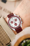 
A close-up shot of a wrist wearing a white Omega x Swatch MoonSwatch "Alaska Project" or "Arctic" watch with red accents, a white dial, and a deep burgundy lizard-patterned watch strap. The arm has light skin and is partially covered by a beige, textured sleeve. The background is softly blurred with light, neutral tones.
