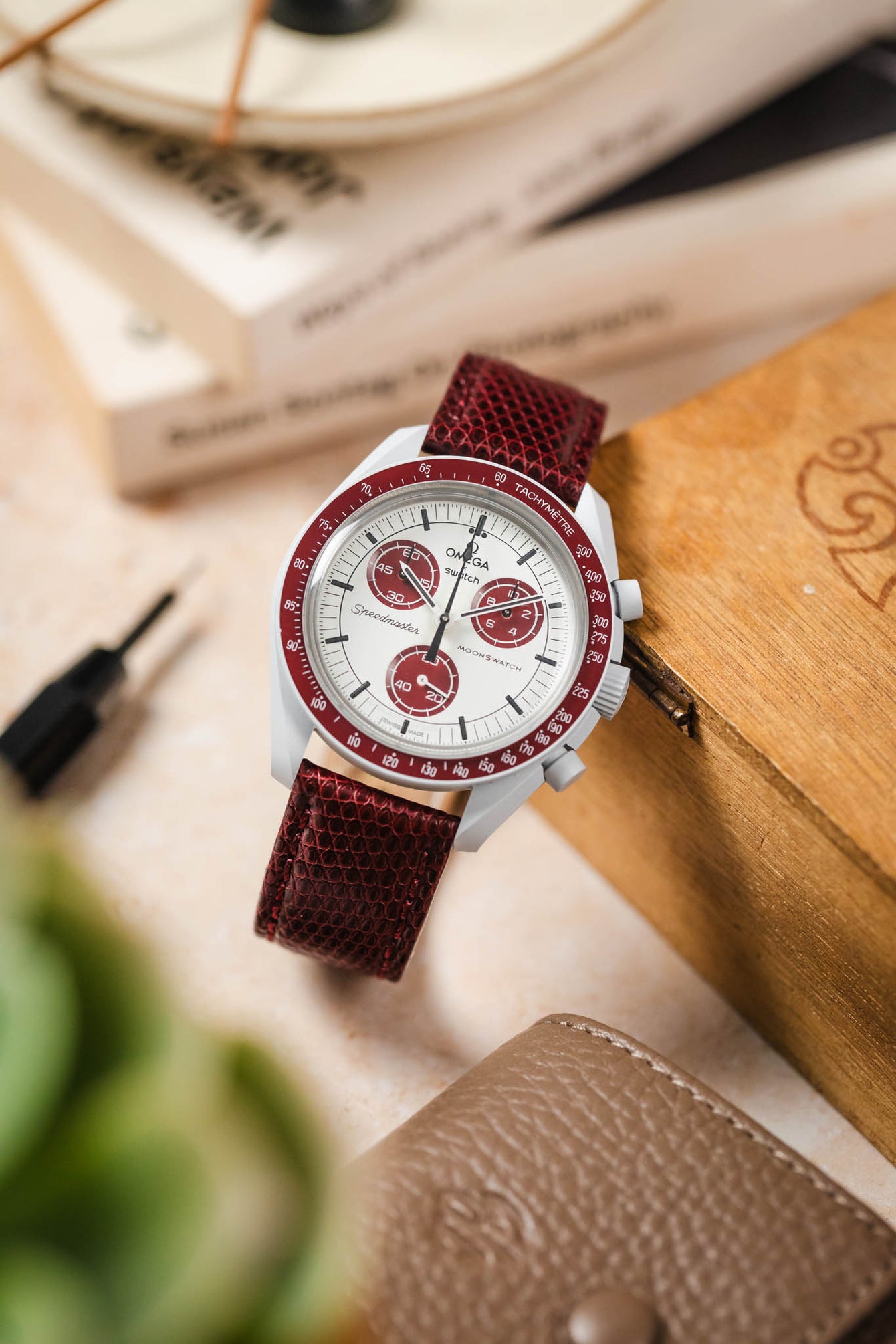 
A top-down, slightly angled shot of a white Omega x Swatch MoonSwatch "Alaska Project" or "Arctic" watch with red accents and a deep burgundy lizard-patterned strap. The watch appears to be floating above a light, textured surface, surrounded by various blurred objects including a wooden box, a brown leather watch pouch, books, and a plant.