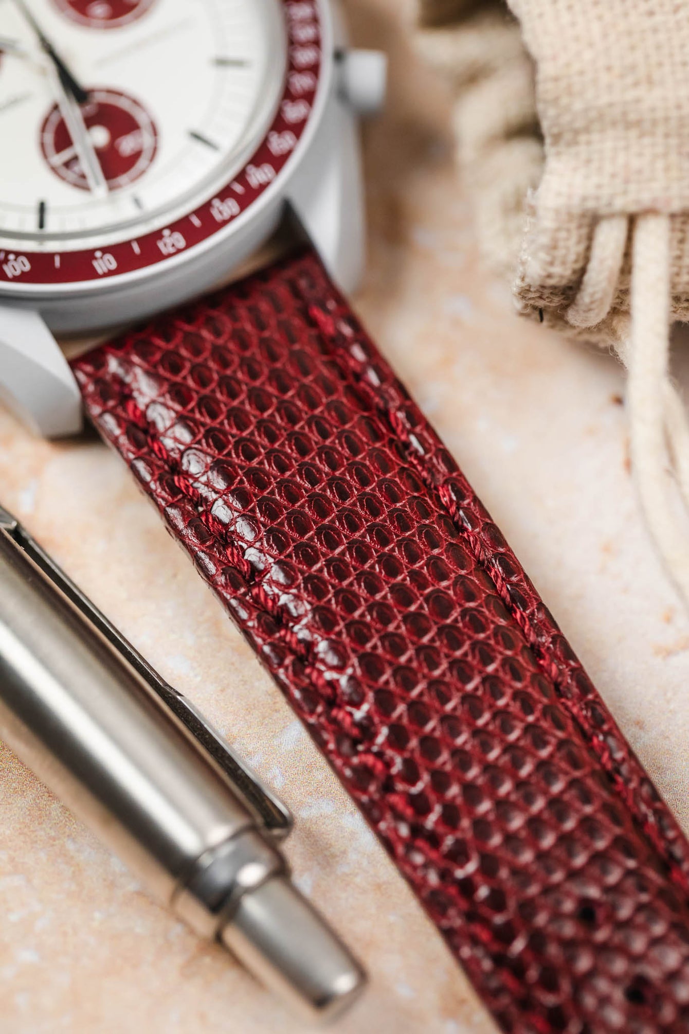A close-up, angled shot focusing on the connection between a white watch case with red accents and a deep burgundy watch strap with a textured, scaly pattern. The strap is visible in the foreground, extending towards the right. A portion of the watch's white dial is visible in the upper left, and a silver pen lies in the lower left. A small, light-colored drawstring bag is blurred in the upper right background. The surface beneath is light and textured.