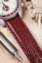 A close-up, angled shot focusing on the connection between a white watch case with red accents and a deep burgundy watch strap with a textured, scaly pattern. The strap is visible in the foreground, extending towards the right. A portion of the watch's white dial is visible in the upper left, and a silver pen lies in the lower left. A small, light-colored drawstring bag is blurred in the upper right background. The surface beneath is light and textured.